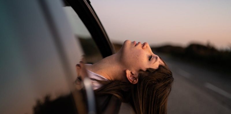 A woman leans out of a car window, enjoying a sunset drive on a quiet road.